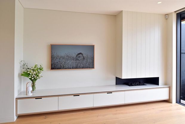 Bright minimalist living room featuring a long built-in lowline cabinetry unit in white, a vertical white-panelled wall and integrated black fireplace niche, pale timber flooring, recessed ceiling lighting, and full-height glazing at the right. A framed artwork and vase with greenery add restrained decorative accents.