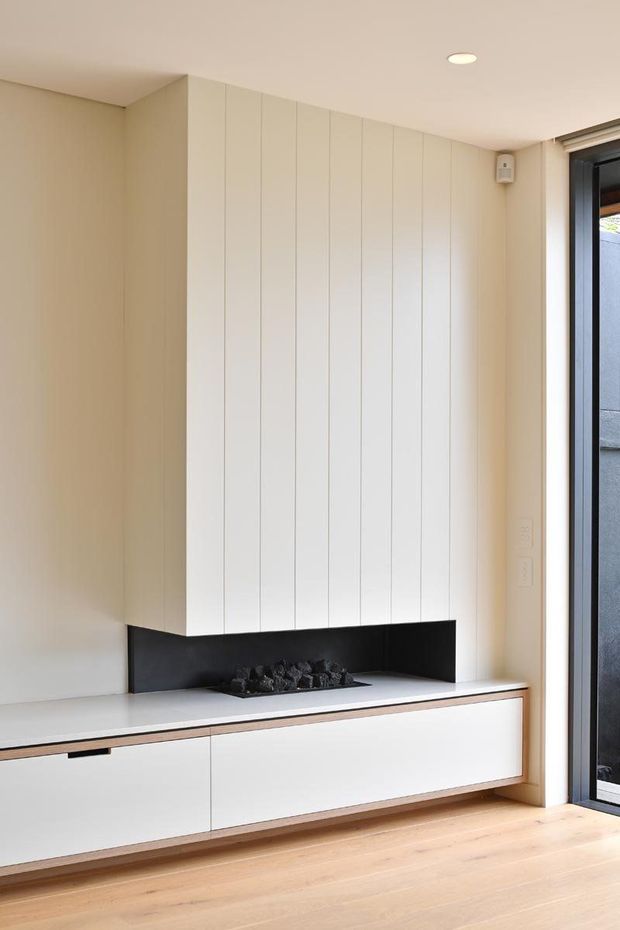 A bright, minimalist living room feature wall with a long white built-in media cabinet and a linear black fireplace. Tall vertical white paneling rises to the ceiling above the fireplace, while pale timber flooring and a large black-framed glazed door add contrast. The palette is warm and neutral with clean contemporary detailing.