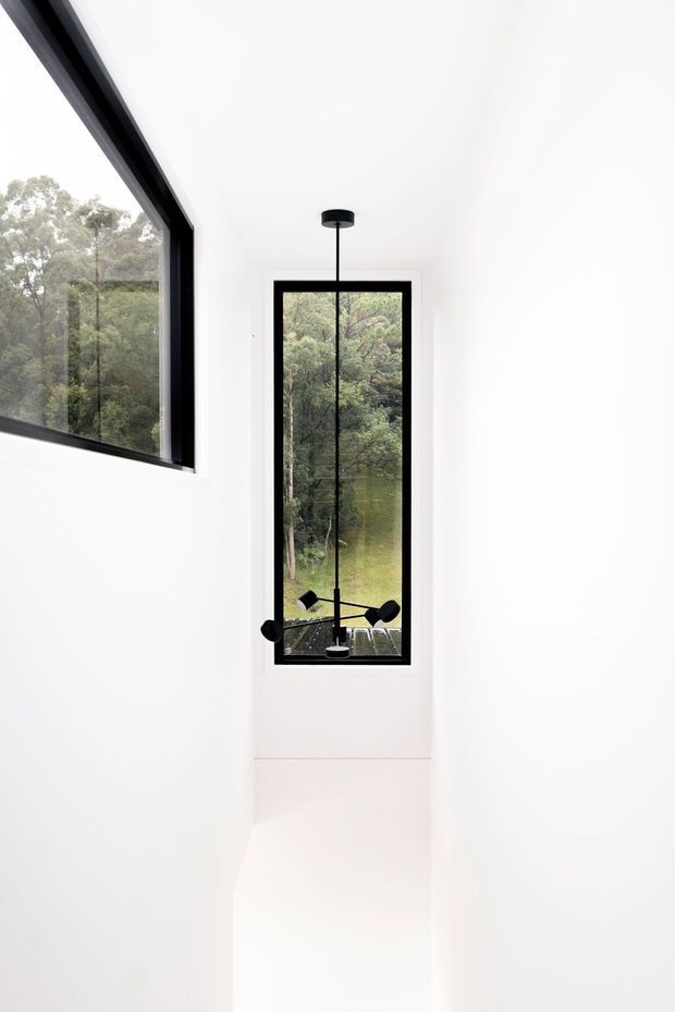 A minimalist white interior passage or stairwell with a tall, narrow black-framed window and a large angled black-framed opening. The glazing looks onto dense green bushland, while a sculptural black pendant light hangs centrally in front of the tall window. The space is bright, sparse and architecturally focused, with smooth white walls and ceiling.