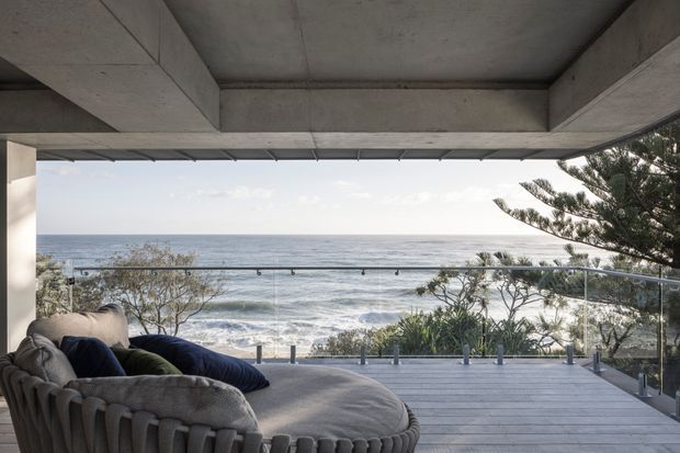 A covered oceanfront outdoor lounge terrace with a large curved upholstered sofa, glass balustrade, timber decking, exposed concrete beams and ceiling, and expansive views over the sea. Coastal vegetation and a Norfolk Island pine frame the outlook.