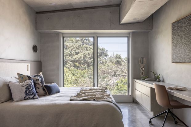 A contemporary bedroom with a low-profile upholstered bed, soft neutral bedding, and decorative cushions. Full-height windows overlook leafy trees, while a built-in light-toned cabinet and compact desk create a work area along the right wall. The room features exposed concrete ceiling and structural detailing, polished grey concrete flooring, and understated decorative accents.