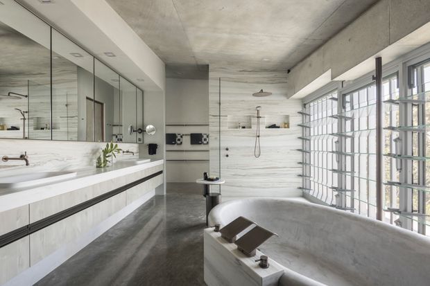 A spacious contemporary bathroom with a large freestanding concrete bathtub in the foreground, a walk-in shower at the rear, floating double vanity with extensive mirrored cabinetry, and expansive industrial-style gridded windows. The room combines pale marble-like wall surfaces, dark polished flooring, exposed concrete ceilings, and restrained black metal fixtures.