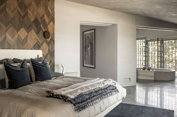 A spacious contemporary bedroom with a large upholstered bed, dark cushions, layered neutral bedding, and a striking geometric timber feature wall behind the bed. The room opens toward a bright adjoining sitting area with a curved upholstered bench, extensive gridded glazing, polished concrete flooring, and a softly textured concrete ceiling. A framed artwork and minimalist architectural detailing complete the refined, modern interior.