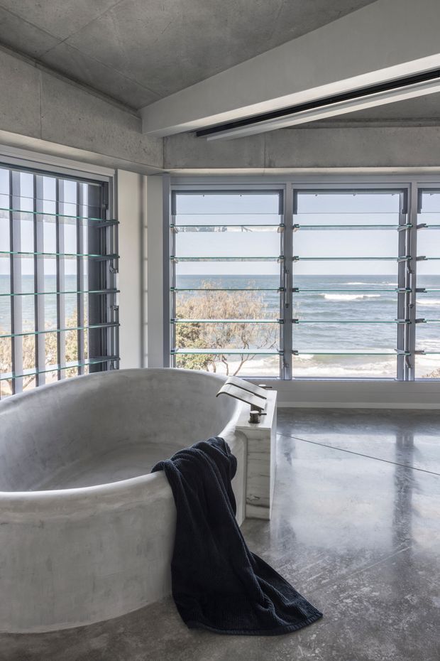 A minimalist concrete bathroom with a freestanding rounded bathtub beside large louvred windows overlooking the ocean. The space features exposed concrete walls and ceiling, a polished concrete floor, pale framed glazing, and a black towel draped over the tub. The expansive coastal view provides strong natural light and a serene atmosphere.
