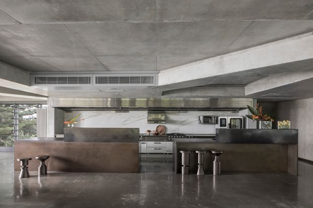 Industrial-style open-plan kitchen with a large dark stone island and peninsula, integrated stainless-steel appliances, marble-look backsplash, concrete ceiling and exposed structural beams. Bar stools and minimal styling create a refined, utilitarian commercial-kitchen feel.