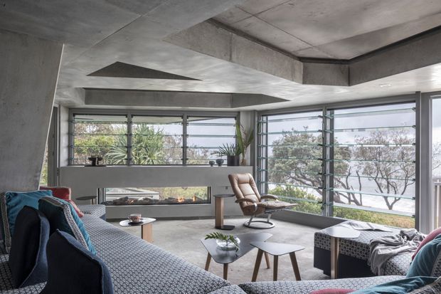 A contemporary living room with exposed raw concrete ceiling beams, columns, and a built-in concrete fireplace. Floor-to-ceiling corner glazing surrounds the room, providing expansive outdoor views. The space includes a large sectional sofa, low timber coffee tables, a leather lounge chair, and restrained modern furnishings in muted grey, blue, and natural tones.