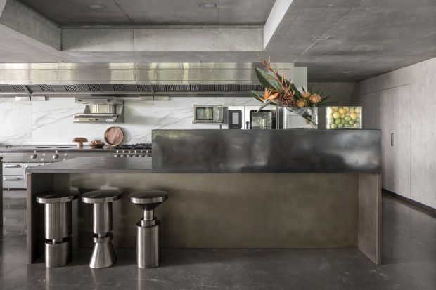 A sophisticated industrial-style kitchen with a large dark metal island and integrated raised counter, three stainless steel bar stools, commercial-grade stainless cooking appliances, white marble-look wall cladding, and an exposed concrete ceiling and floor. A linear ventilation hood and open shelving reinforce the professional kitchen character, while floral arrangements and fruit add warmth.