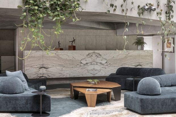 A sophisticated contemporary living room with deep charcoal and blue-grey sofas arranged around a sculptural timber coffee table. A dramatic veined stone feature wall or fireplace surround spans the rear of the room, set beneath a concrete-toned ceiling beam. Trailing indoor plants soften the restrained palette, while sculptural decor and layered rugs add texture.