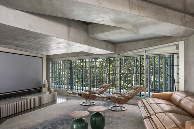A spacious, minimalist media and living room with exposed raw concrete ceilings and walls, extensive floor-to-ceiling glazing with vertical metal security screens, a large projection screen, leather lounge seating, and sculptural lounge chairs. The space has a refined industrial character and overlooks dense greenery.