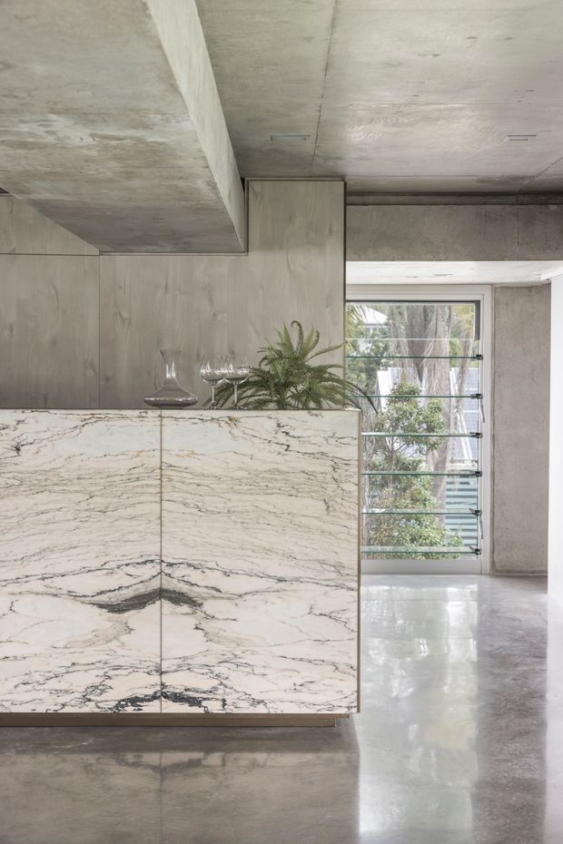 A refined contemporary interior featuring a large marble-clad island or built-in cabinet with dramatic veining, set against raw concrete walls, ceiling, and polished concrete flooring. A small plant and glassware sit on the stone surface, while a louvred window provides natural light and views of greenery.