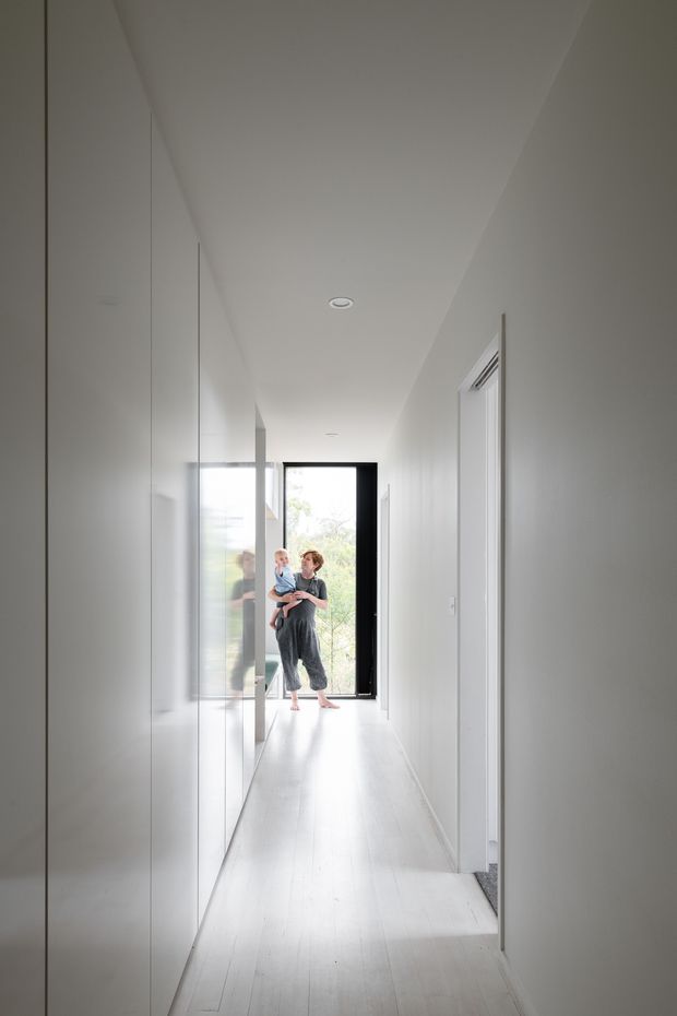 A bright, narrow residential hallway with white walls, a white linear timber floor, full-height built-in cabinetry on the left, and several white doorways on the right. A black-framed glazed entry door at the far end opens to greenery outside, with an adult holding a child standing in the doorway. The space is minimalist, clean, and strongly illuminated by natural light.