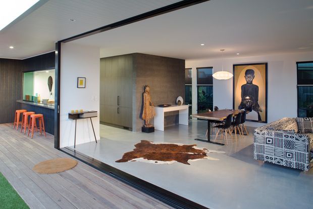 Open-plan contemporary interior viewed through broad black-framed glazing, combining a kitchen, dining area and living space. The room has pale concrete-look flooring, dark timber wall panelling, extensive built-in cabinetry, a white island, a large dining table, patterned lounge seating, recessed lighting and a sculptural pendant. A timber deck and outdoor bar area are visible beyond the glazing.