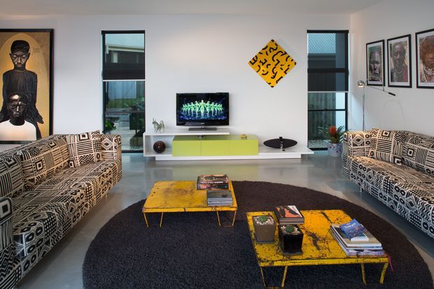 A stylish modern living room with two large patterned sofas facing a wall-mounted television and low lime-green media console. Two distressed yellow coffee tables sit on a large dark shag rug, while bold contemporary artwork and framed portraits decorate the white walls. Tall narrow windows provide natural light and reveal an urban residential setting.