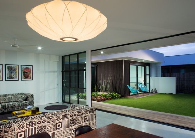 A contemporary open-plan living and dining area opens through extensive black-framed glazing to a landscaped courtyard. The interior features a large patterned sectional sofa, a dark timber dining table, white walls, polished concrete flooring, and a sculptural pendant light. Outside, a compact lawn, planted garden bed, timber decking, and a flat-roofed pavilion with dark vertical cladding are visible.