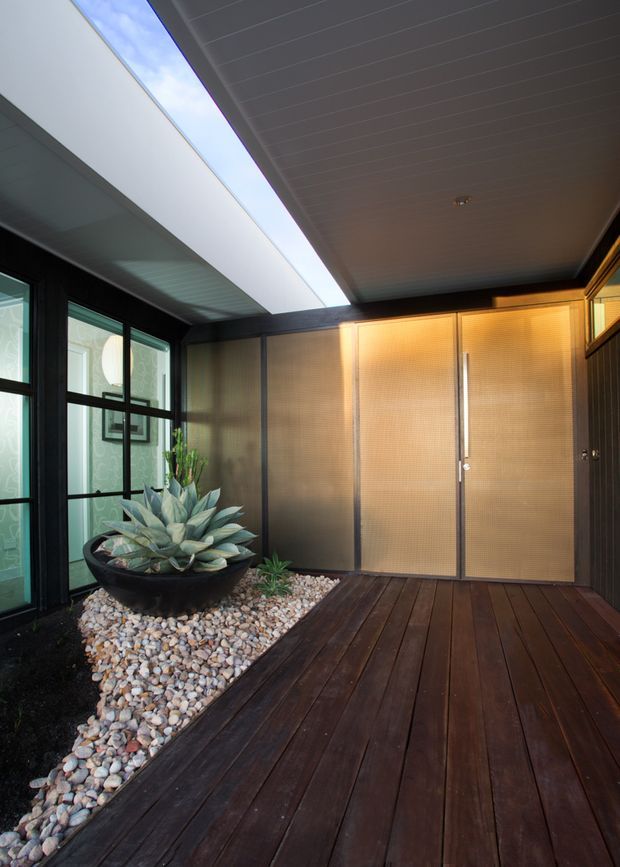 A covered residential entry courtyard with dark timber decking, a pebble garden bed containing a large sculptural planter, black-framed glazing, and illuminated textured double entry doors. A long linear skylight brings daylight into the sheltered space, creating a refined modern entrance.