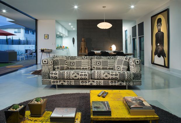 A contemporary open-plan living room with a bold black-and-white geometric patterned sofa facing a pair of distressed yellow coffee tables. The space has polished concrete flooring, a dark feature wall with a fireplace and pendant light, recessed ceiling lighting, large sliding glass doors opening to an outdoor patio, and a striking framed artwork on the right wall. A kitchen or dining zone is visible beyond the living area.