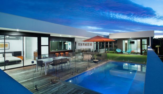 Wide twilight exterior view of a contemporary single-storey residence arranged around a timber pool deck. The composition shows extensive floor-to-ceiling glazing, flat rooflines, an outdoor dining and lounge area with umbrella, and a rectangular swimming pool reflecting the illuminated house. Interior living spaces are visible through the glazing.