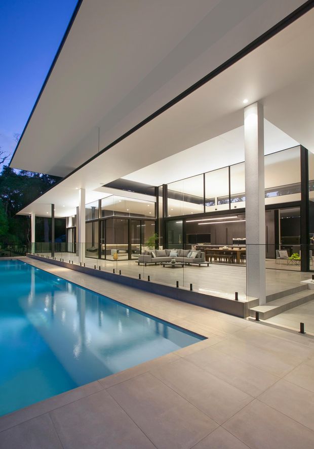 Contemporary luxury residence viewed from the pool terrace at dusk, featuring a broad flat roof, extensive floor-to-ceiling glazing, slender dark frames, white structural columns, and an open-plan living, dining, and kitchen area illuminated inside. The foreground is dominated by a long swimming pool and pale paved terrace.
