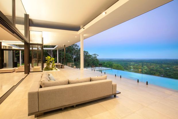 A spacious contemporary covered outdoor living terrace overlooking a lush landscape, with a large beige sectional sofa, pale tiled flooring, slim white columns, extensive glazing, and an infinity-edge swimming pool. The open-plan space has a broad cantilevered roof and integrated warm lighting, creating a refined indoor–outdoor connection.