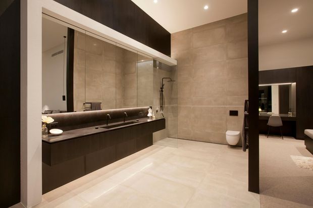 Contemporary bathroom with a large mirrored double vanity, dark floating cabinetry, integrated basins, full-height glass shower enclosure, wall-mounted toilet, and large-format beige stone-look wall and floor tiles. A bedroom or dressing area is visible through the open doorway at right.