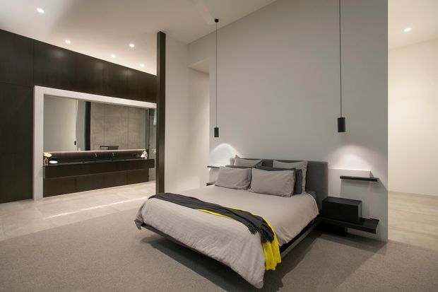 Contemporary bedroom with a low upholstered bed, layered neutral bedding, dark bedside furniture, and minimalist pendant lights. A large open ensuite bathroom with dark built-in cabinetry and a freestanding bath is visible behind the bedroom, while recessed ceiling lighting and soft grey finishes create a refined, calm atmosphere.