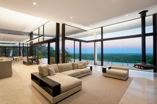 A spacious, refined open-plan living room with floor-to-ceiling dark-framed glazing overlooking a broad coastal or ocean-facing landscape. The room features a large neutral sectional sofa, a matching ottoman, a low black coffee table, pale tiled flooring, recessed ceiling lighting, and a striking suspended black fireplace. A dining area and kitchen are visible in the background.