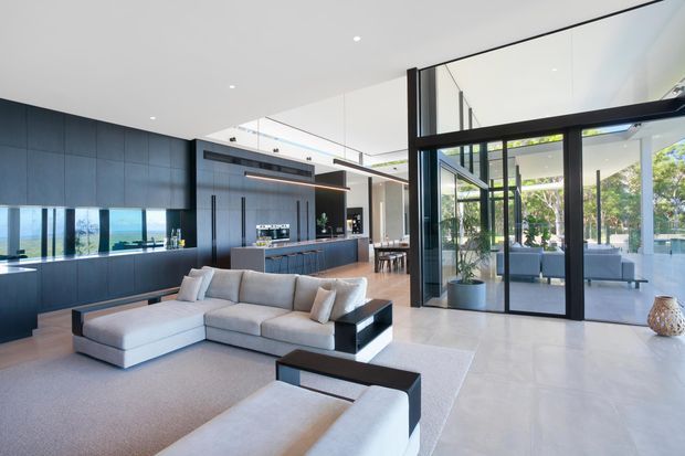 Open-plan contemporary living space with a large light sectional sofa in the foreground, a dark blue-grey kitchen with extensive built-in cabinetry and island behind it, and full-height black-framed glass doors opening to a bright outdoor terrace. The room has a minimalist white ceiling with recessed lighting, pale flooring, and strong indoor-outdoor visual connection.