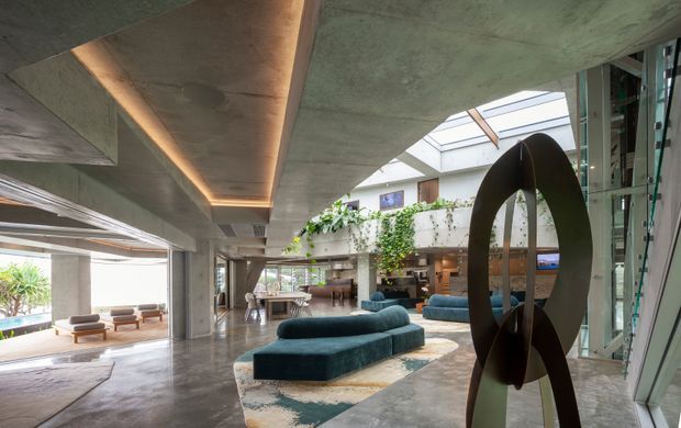 A spacious, high-end contemporary interior centered on a dramatic multi-level living and lounge area. The space features exposed concrete ceilings and columns, polished concrete flooring, extensive glazing, a large skylight, integrated warm lighting, indoor greenery, sculptural artwork, and low teal seating arranged around patterned rugs. A dining area and additional lounge spaces are visible in the open-plan interior.