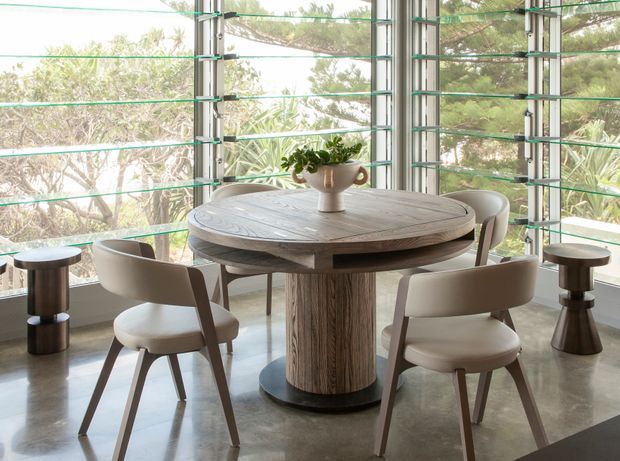 Bright, airy dining area with a round sculptural timber dining table and upholstered dining chairs arranged beside expansive corner glazing. Horizontal louvres and slim metal window framing overlook leafy trees, while small side tables and a simple ceramic vase add restrained styling. The polished concrete floor and abundant natural light create a refined contemporary setting.