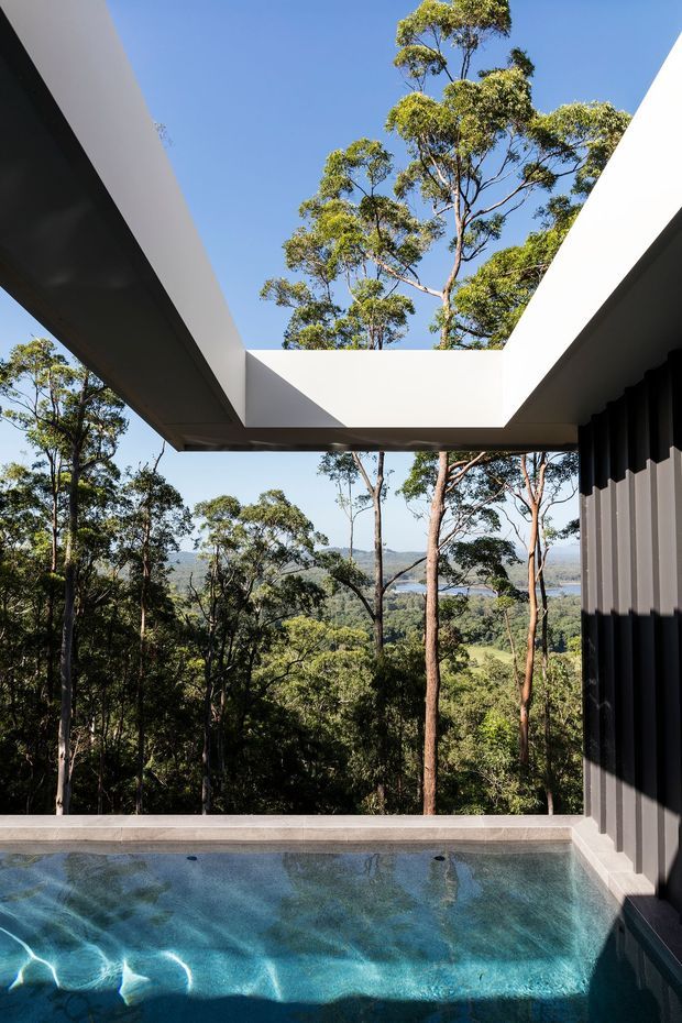 A contemporary outdoor pool terrace set within a wooded, elevated landscape. The compact rectangular pool sits beneath a dramatic white flat-roof canopy with a large open skylight framing the sky and surrounding trees. Dark vertical wall cladding contrasts with the white roof structure, while a distant valley and water view are visible through the open sides.