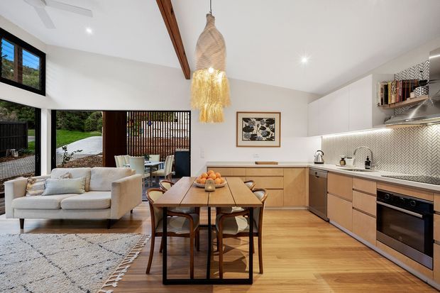 Open-plan kitchen and dining area adjoining a bright living space. The room features white walls and ceiling, warm timber flooring, light timber kitchen cabinetry, a pale stone or composite benchtop, and a patterned tile splashback. Black-framed glazing opens toward a leafy outdoor setting, while a timber ceiling beam and woven pendant light add warmth. A dining table, upholstered chairs, sofa, rug, integrated oven, and open shelving are visible.