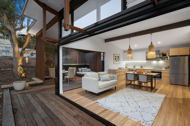 A contemporary open-plan living, dining and kitchen area viewed from a timber deck through expansive black-framed glazing. The interior features pale timber flooring, white walls, exposed dark timber beams, a compact kitchen with timber cabinetry and a tiled backsplash, pendant lights over a dining island, and a white sofa grouping. A planted courtyard and vertical timber screening are visible outside.