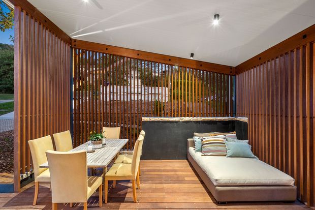 A covered outdoor entertaining area with warm timber slat screening, a white ceiling with recessed-style spotlights, timber decking, a dining table with chairs, and a large outdoor sectional lounge. The partially open slatted walls provide views to surrounding greenery and a driveway beyond.