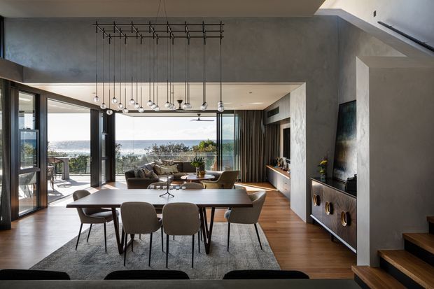 A refined open-plan dining and living area with warm timber flooring, a large dining table and upholstered chairs in the foreground, and a lounge seating area beyond. Full-height glazed doors open toward a coastal ocean view and balcony. The space features textured grey concrete-look walls, dark-framed glazing, timber-lined details, a contemporary linear pendant installation, built-in media cabinetry, and a stairway at right.