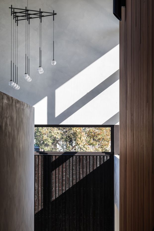 A narrow, double-height interior passage or entry space with dramatic sunlight casting strong geometric shadows across smooth plaster walls. A sculptural black linear pendant with multiple suspended glass bulbs hangs from the ceiling. Dark vertical timber or metal screening sits beneath a large horizontal window, while a warm vertical timber-clad wall frames the right side. The image has a refined, modern architectural character with strong contrast and carefully controlled natural light.