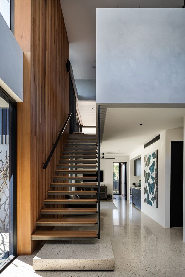 A bright contemporary interior centered on a floating timber staircase with slim black metal handrails and open risers. The staircase is set against a full-height vertical timber-panelled wall and rises beside a large white plastered volume. Polished terrazzo flooring continues through the open-plan space, with white walls, a black-framed glazed door, recessed lighting, and glimpses of living and dining areas beyond. The composition highlights the stairway, double-height architectural volume, and contrasting timber, white, and dark metal finishes.