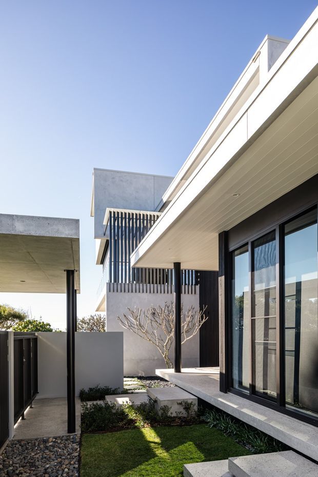 Exterior architectural photograph of a contemporary multi-level residence viewed from a landscaped courtyard. The composition highlights clean white rendered and concrete forms, deep roof overhangs with a pale lined soffit, dark metal posts and window frames, vertical screening, and large glazed doors opening onto a raised terrace. Stepping paths, gravel beds, lawn, and sculptural planting create a minimalist courtyard setting under a clear blue sky.