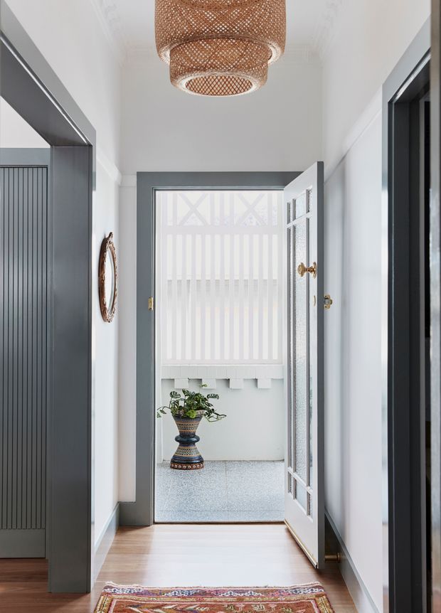 Bright, narrow residential entry hall leading to an open glazed front door and a small exterior threshold area. The hallway has white walls and ceiling, dark charcoal-grey door and wall trim, warm timber flooring, a patterned runner, and a woven rattan pendant light. A decorative planter is visible beyond the doorway, while dark built-in joinery or a doorway frames the left side.