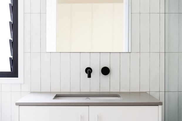 A minimalist bathroom vanity with a pale grey engineered stone benchtop, integrated rectangular basin, white cabinetry, and matte black wall-mounted tapware. Vertical white tiles with dark grout form the feature wall, accompanied by a large frameless mirror and a black-framed window.