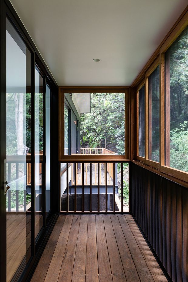 A narrow enclosed timber-decked passage or balcony with extensive glazing, dark metal-framed doors on the left, warm timber window frames on the right, and a timber-and-metal balustrade at the end. Dense greenery and an adjacent deck are visible outside. The space has a simple white ceiling with a recessed light and a strong indoor-outdoor connection.