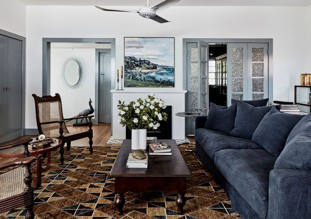 A refined living room with a deep navy-blue sectional sofa, dark wood coffee table, patterned geometric rug, and traditional cane armchairs. White walls, grey-framed openings, decorative patterned glass doors, a fireplace mantel with artwork and flowers, and a ceiling fan create a layered transitional interior with coastal artwork and classic furnishings.