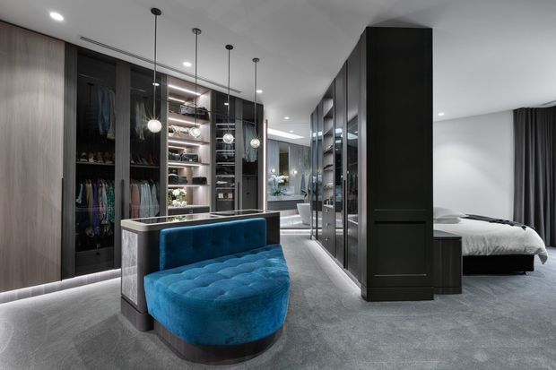 A luxurious walk-in wardrobe and dressing area with floor-to-ceiling dark cabinetry, glass-fronted clothing storage, illuminated shelving, pendant lights, and a curved tufted blue upholstered bench. A bedroom with a neatly made bed is visible beyond the central partition. The space has grey carpeting, dark timber cabinetry, and a refined contemporary fit-out.
