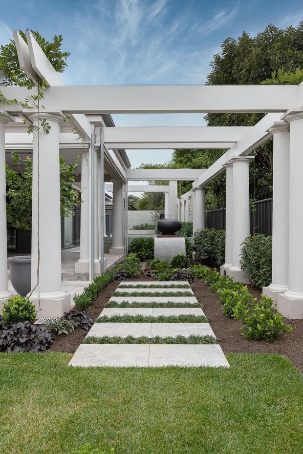 A landscaped courtyard walkway beside a modern home, featuring large pale stepping slabs set through lawn and garden beds. White painted columns and a rectangular pergola create a covered passage, with glass walls and dark metal fencing alongside clipped shrubs and leafy planting. A dark sculptural object sits on a pale pedestal at the far end. The scene is bright, refined, and symmetrical, with a contemporary-classical character.