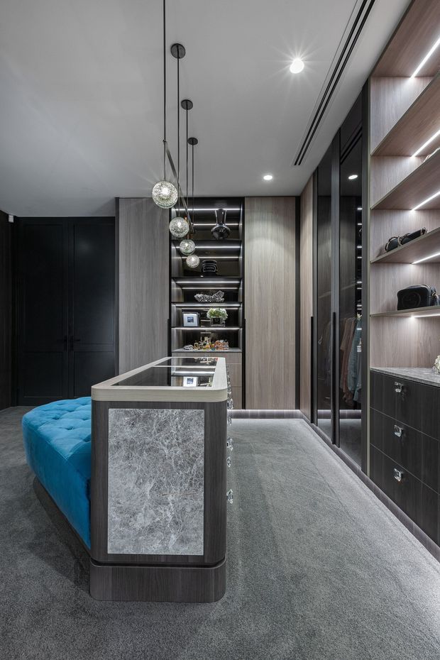 A luxurious walk-in wardrobe with dark timber cabinetry, full-height mirrored wardrobe doors, illuminated open shelving, plush grey carpet, and a central island featuring a marble-look panel and glass top. A vivid blue upholstered bench curves around the island, while pendant globe lights add a refined decorative element.