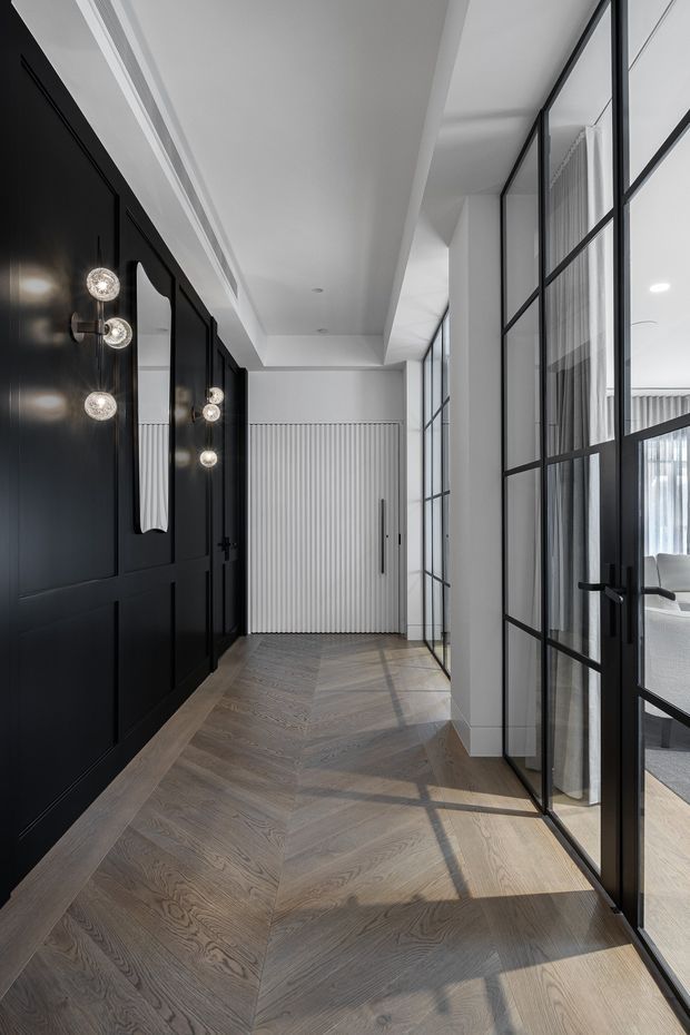 A refined contemporary hallway with a warm grey-brown herringbone timber floor, white walls and ceiling, and dramatic black built-in wall panelling. Globe wall lights are mounted along the dark joinery, while black-framed glazed partitions and doors bring natural light into the corridor and reveal a softly furnished room beyond. A vertically ribbed white built-in door anchors the far end.