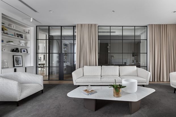 Refined contemporary living room with a large white sectional sofa, sculptural white coffee table, dark grey carpet, full-height beige curtains, and black-framed glazed partitions opening to a study or workspace. Built-in display shelving lines the left wall, while recessed and linear ceiling lighting contribute to the clean, sophisticated interior.