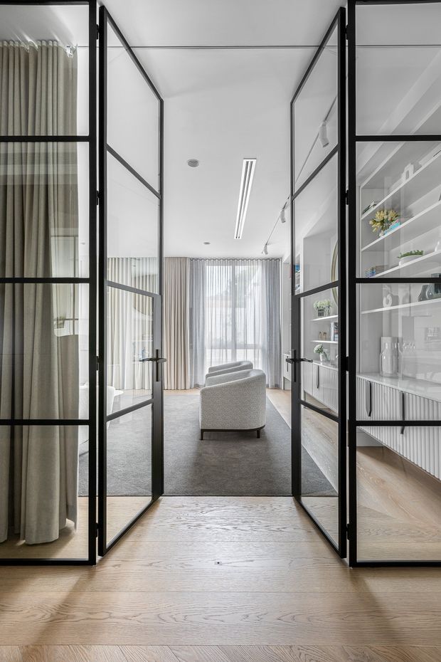 View through open black-framed glazed double doors into a refined living room. The room features pale timber flooring, a large neutral rug, a light upholstered lounge chair, full-height sheer and draped curtains, built-in white display shelving and cabinetry, and slim linear ceiling lighting. The overall palette is bright and neutral with strong black-framed glazing and doors.