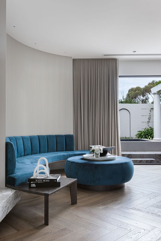 A refined contemporary living room with a curved, vertically fluted feature wall, a deep blue upholstered banquette and matching rounded ottoman, a dark wood coffee table, and full-height taupe curtains. Pale herringbone timber flooring and a large opening to a planted outdoor courtyard create a bright, sophisticated interior.