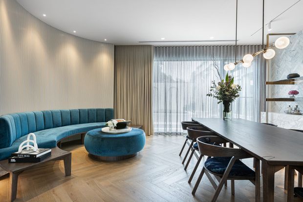 A refined open-plan living and dining room with a curved teal upholstered banquette, round ottoman, dark timber coffee table, long dining table and upholstered dining chairs. Vertical fluted wall panelling, sheer curtains, pale timber flooring, a marble-look feature wall with open shelving, and a sculptural globe pendant create a contemporary, softly luxurious interior.