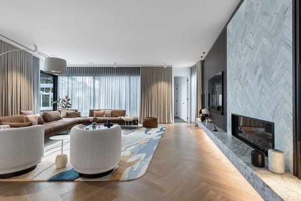 A refined contemporary living room with a large sectional sofa, rounded upholstered lounge chairs, coffee tables, and a patterned area rug. Full-height sheer curtains and drapes line the glazed wall, while a dramatic marble-look herringbone feature wall incorporates a television and long linear fireplace. Warm timber herringbone flooring, dark vertical wall panelling, and layered neutral furnishings create a polished, inviting interior.